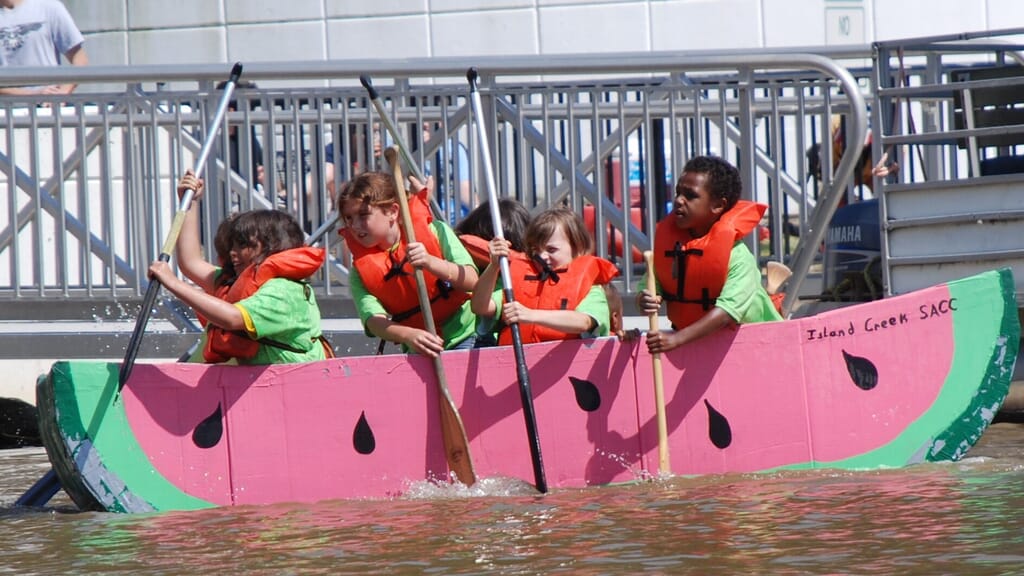 Competitors racing cardboard boats on water, participating in cardboard boat racing, one of the strangest sports in the USA.