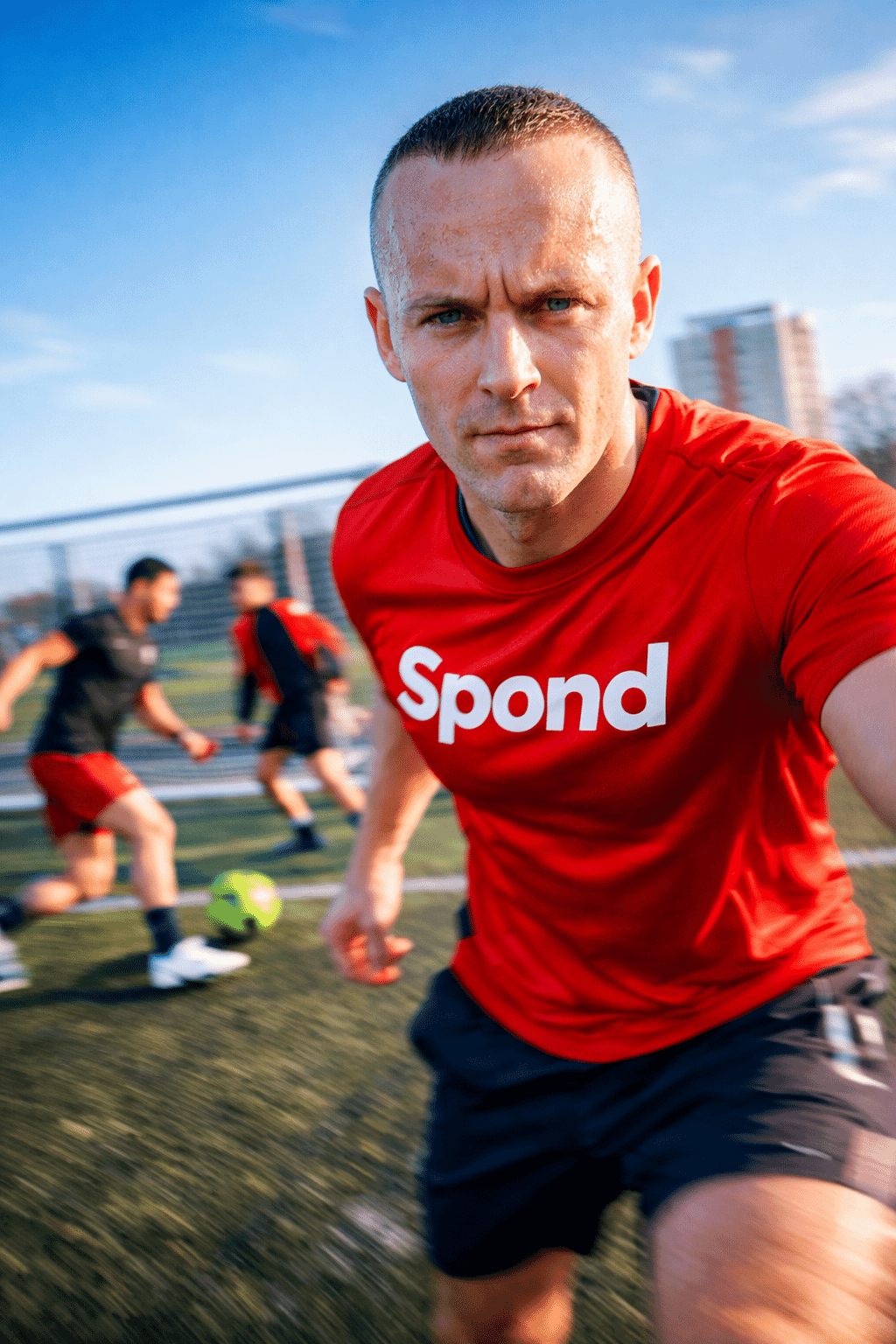 Soccer trainer in a red Spond T-shirt leaning toward the camera on a football field while players train behind him with motion blur.